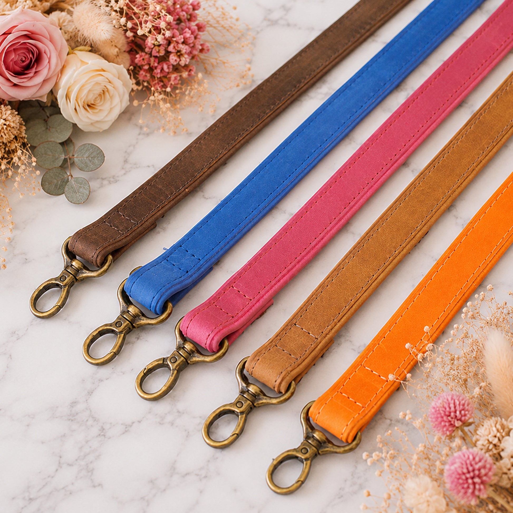 Five colorful leather straps with metal hooks on a marble surface with flowers.