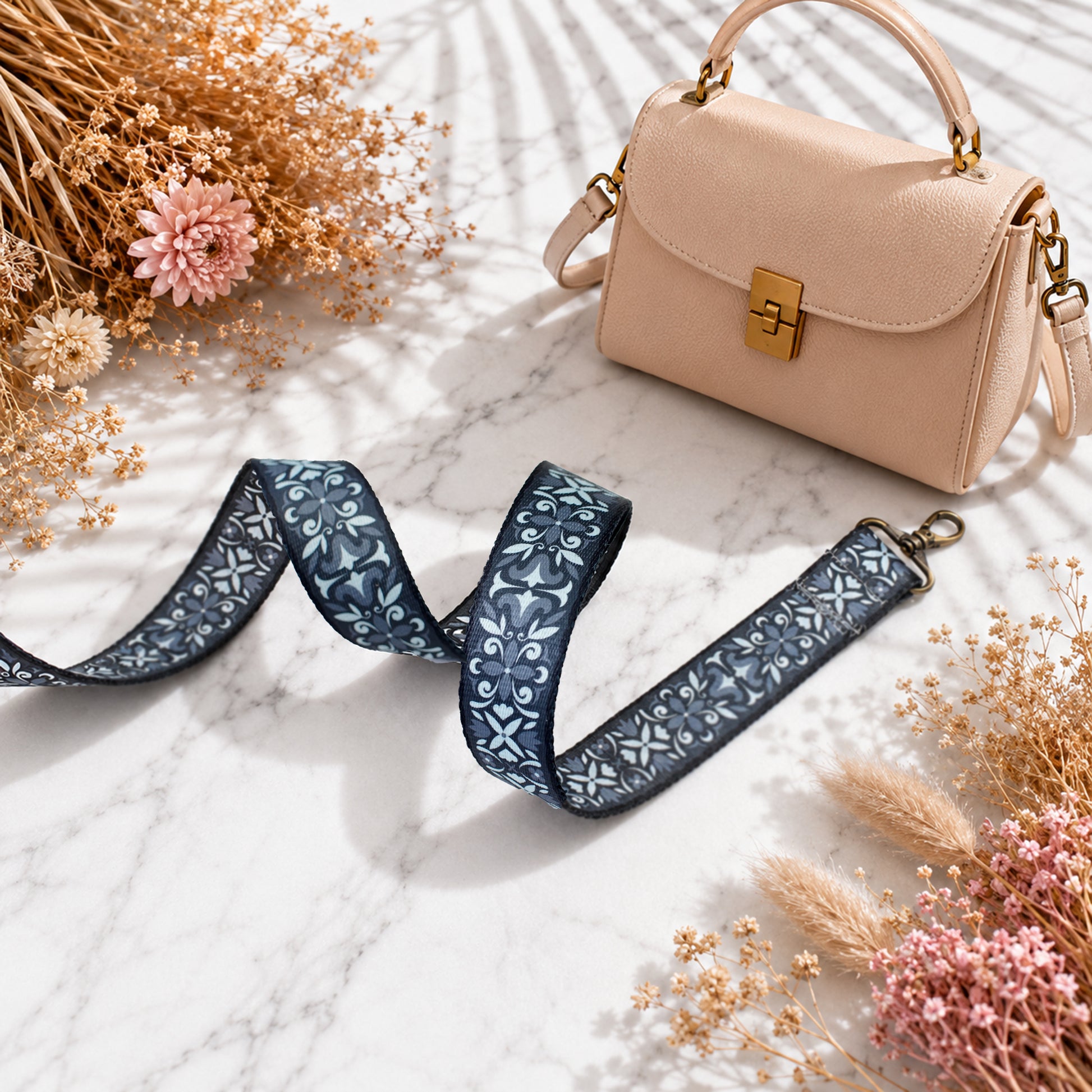 Beige handbag with a floral strap on a marble surface with dried flowers.