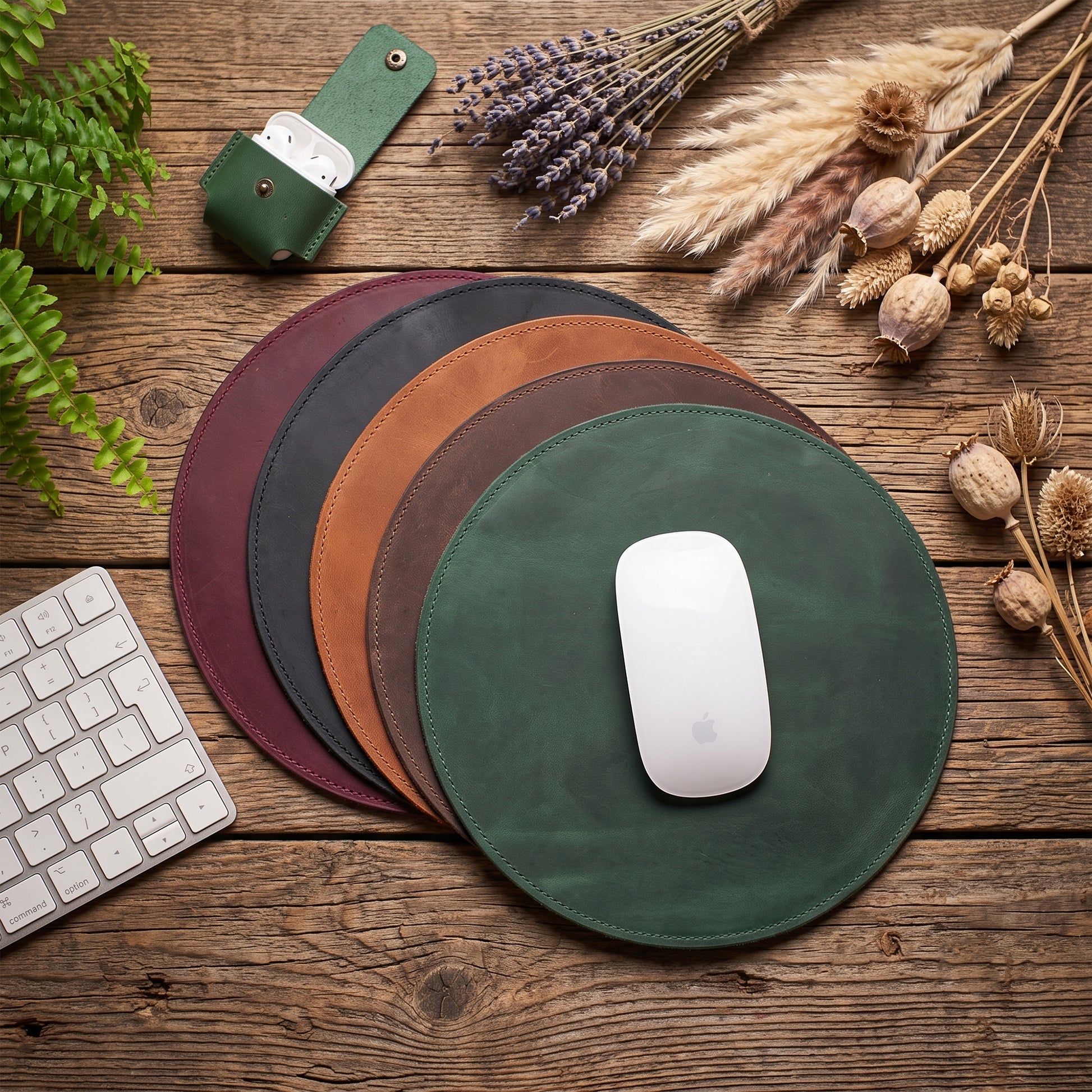 Four round leather mouse pads in green, brown, tan, and burgundy are arranged in a fan shape on a rustic wooden desk, with a white mouse on the front pad. A keyboard, a green leather card holder, dried lavender, and dried grasses are styled around them.