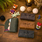 Three personalized leather pouches or organizer cases arranged on a wooden tabletop, alongside yarn balls, a small stuffed tiger, a crochet bear, a lantern, and other cozy handmade props. The pouches are shown in dark green, brown, and black, each with a different embroidered name: Rachel, Ann, and Kelly.