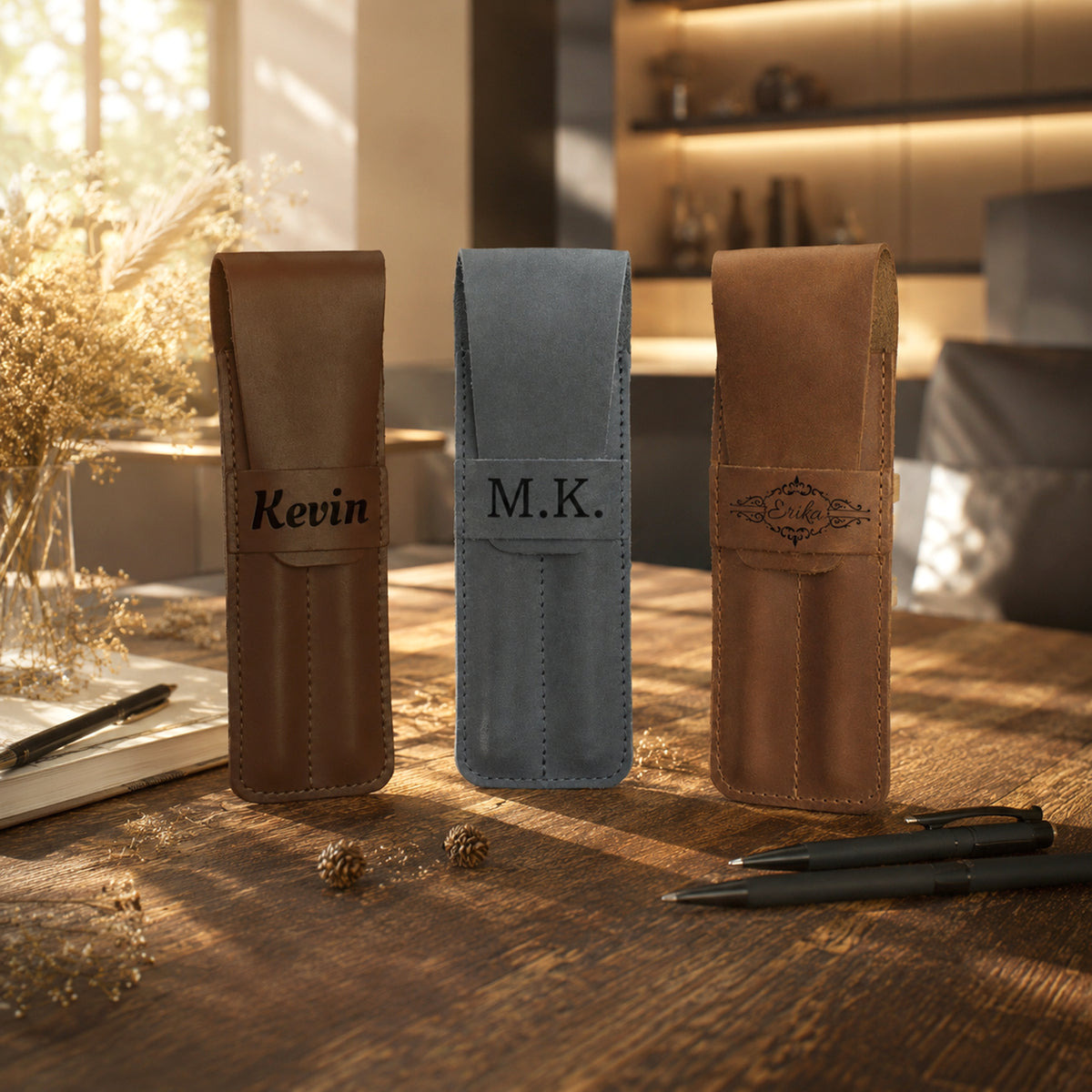 Three personalized leather pen sleeves standing on a wooden table in a warm, sunlit room. The left sleeve is brown with the name 'Kevin' in black script, the center sleeve is light blue with 'M.
