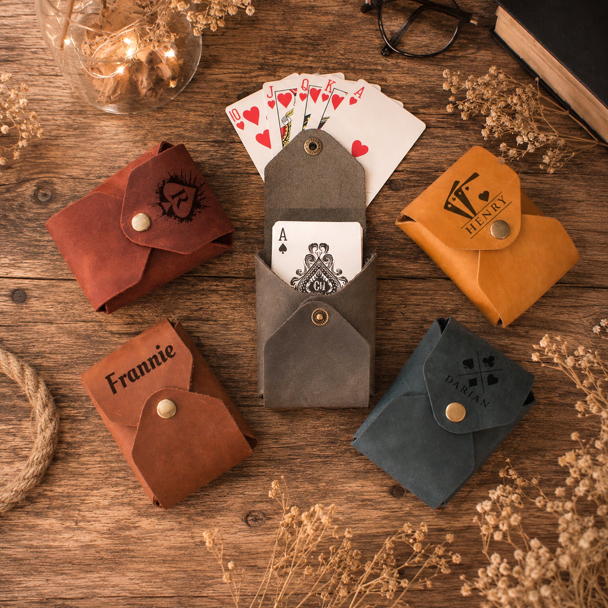 Five small leather card sleeves or wallet-style holders in different colors are arranged on a wooden surface with playing cards, glasses, dried flowers, and other styled props. The center item is open and holds an Ace of Spades card.