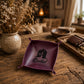 A square purple leather tray with small loop handles at the corners, featuring a black printed house silhouette and the name 'Rachel' on the bottom center. The tray is displayed on a rustic wooden table in a cozy, warm-toned room setting with a vase of dried flowers and other decorative items in the background.