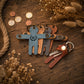 A set of four wooden keychains in blue, brown, tan, and gray, arranged on a rustic wooden surface with dried flowers, rope, and coins. One keychain is shown with keys attached.