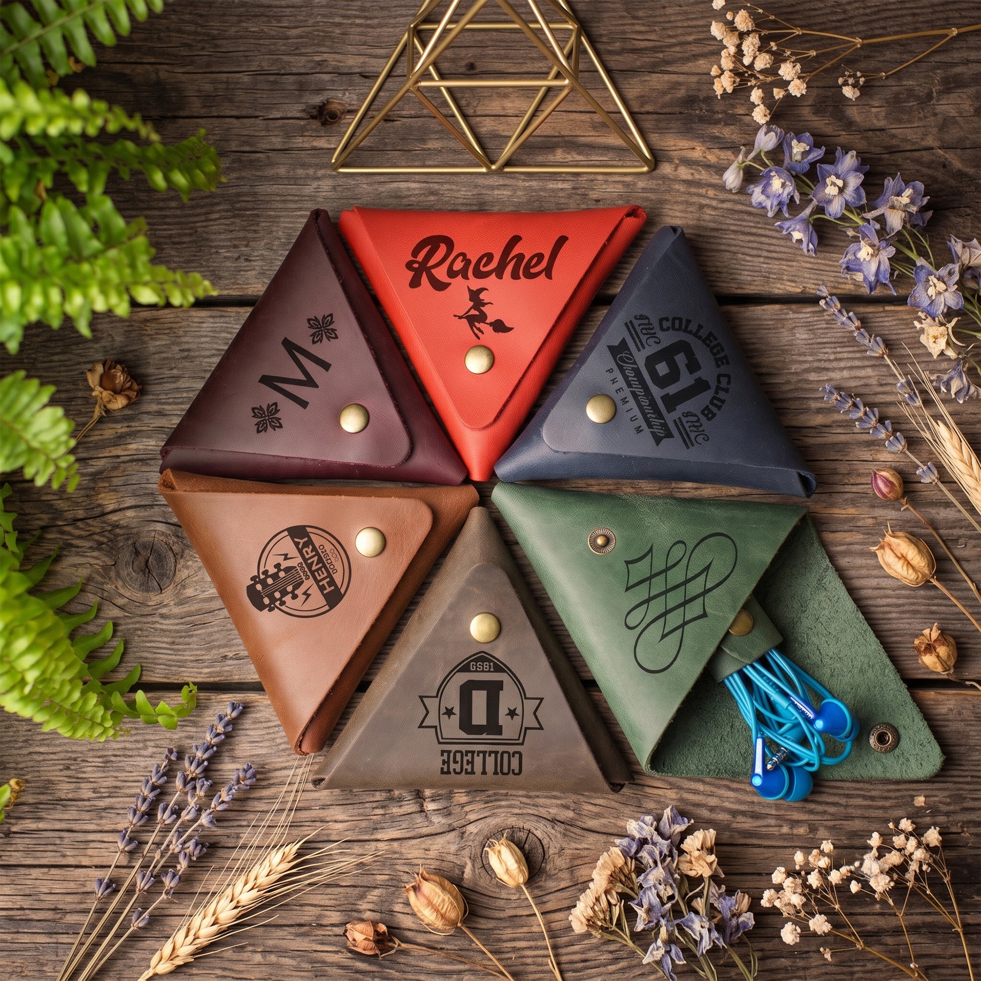 Six triangular leather pouches arranged in a circle on a rustic wooden surface, each in a different color and printed with a monogram or logo. Greenery and dried flowers are styled around the products.