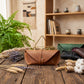 A brown leather envelope-style clutch or case sits on a rustic wooden table, surrounded by dried wheat, lavender, cotton, and other leather cases in green, black, and brown. A gold geometric stand is behind the main case, with a bookshelf and greenery in the softly lit background.