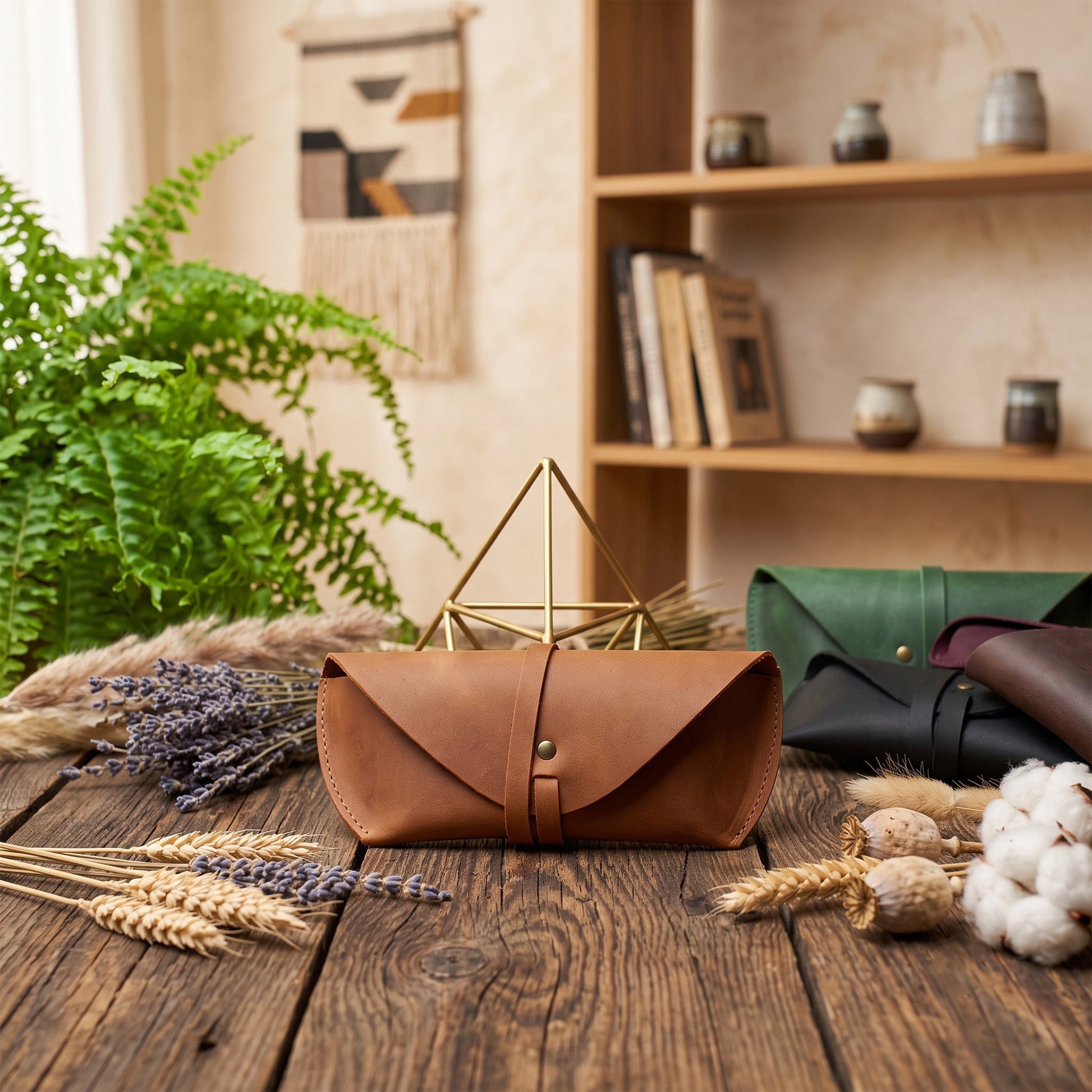A brown leather envelope-style clutch or case sits on a rustic wooden table, surrounded by dried wheat, lavender, cotton, and other leather cases in green, black, and brown. A gold geometric stand is behind the main case, with a bookshelf and greenery in the softly lit background.