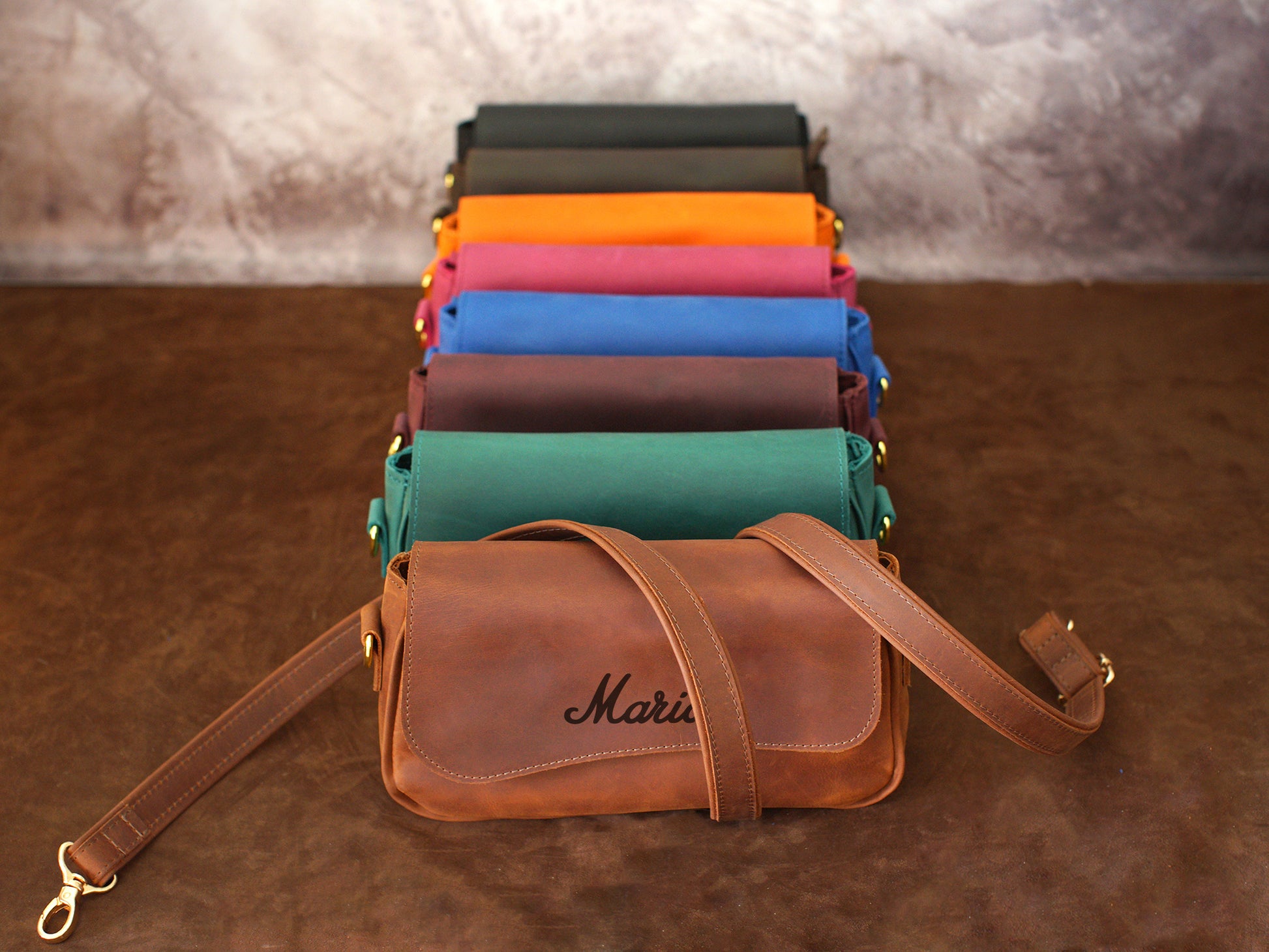 Brown leather bag with 'Maria' branding in front of colorful fabric rolls on a brown surface.