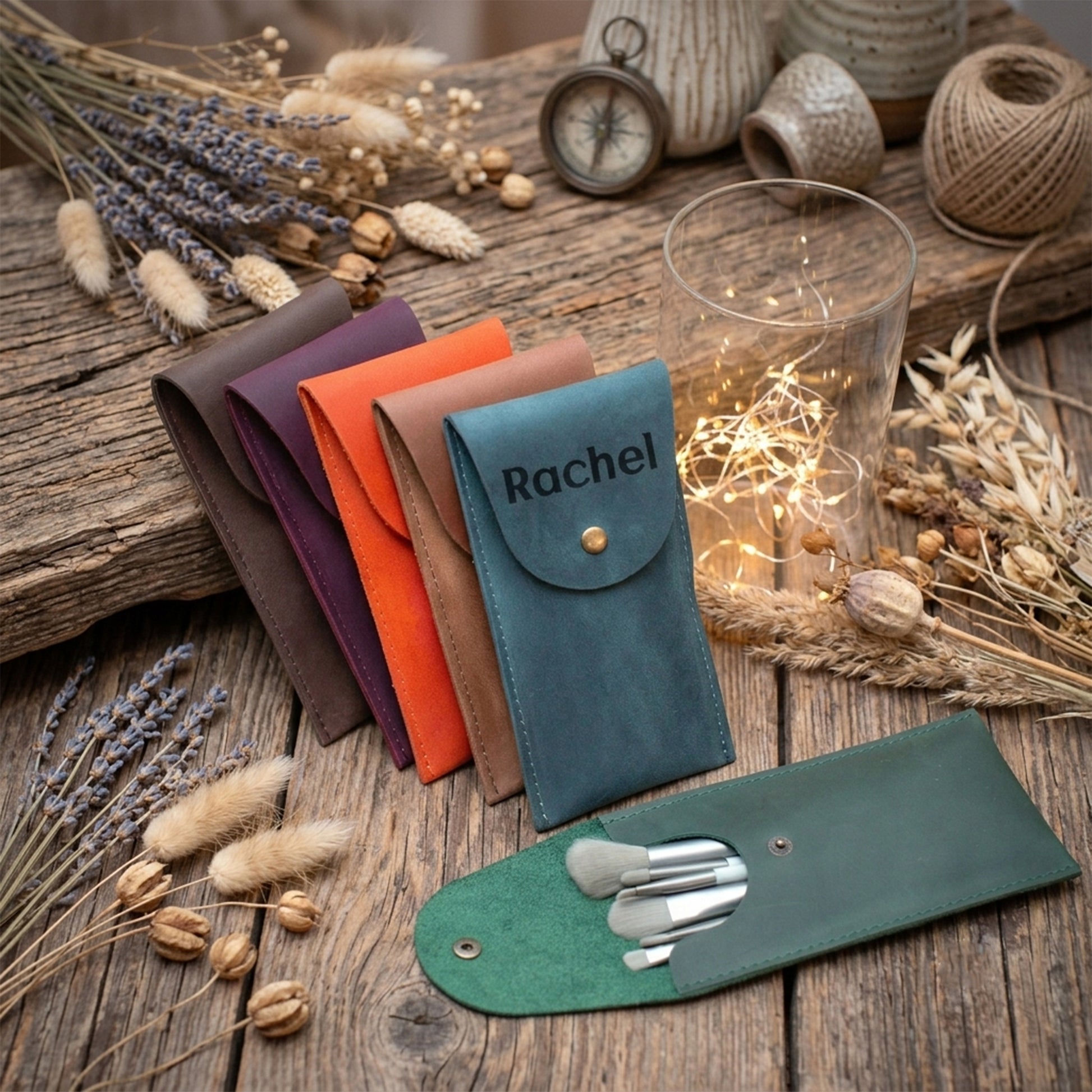 A set of personalized leather pouches in multiple colors, including teal (personalized with 'Rachel'), green, orange, brown, and purple, displayed on a rustic wooden table with dried flowers, a glass candle holder with fairy lights, and makeup brushes.