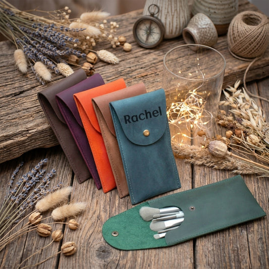 A set of personalized leather pouches in multiple colors, including teal (personalized with 'Rachel'), green, orange, brown, and purple, displayed on a rustic wooden table with dried flowers, a glass candle holder with fairy lights, and makeup brushes.