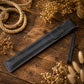 A black leather sheath or sleeve with a matching small strap, laid diagonally on a rustic wooden surface surrounded by dried flowers and rope.