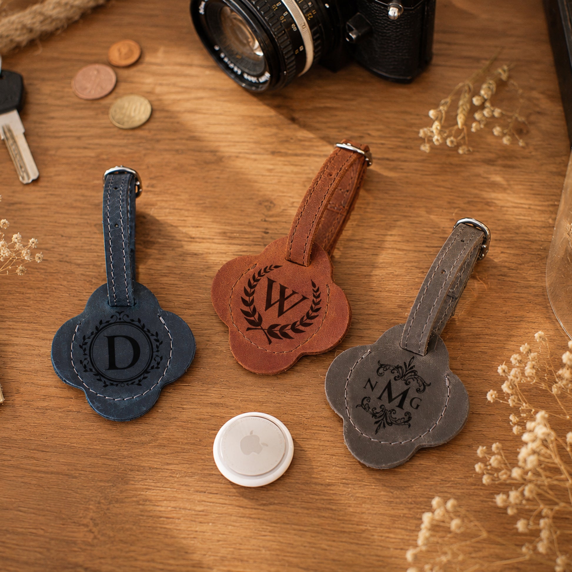 Three personalized leather luggage tags in navy blue, brown, and gray, each shaped like a flower with a stitched loop and monogrammed center, arranged on a wooden tabletop with a camera, coins, and dried flowers as props.