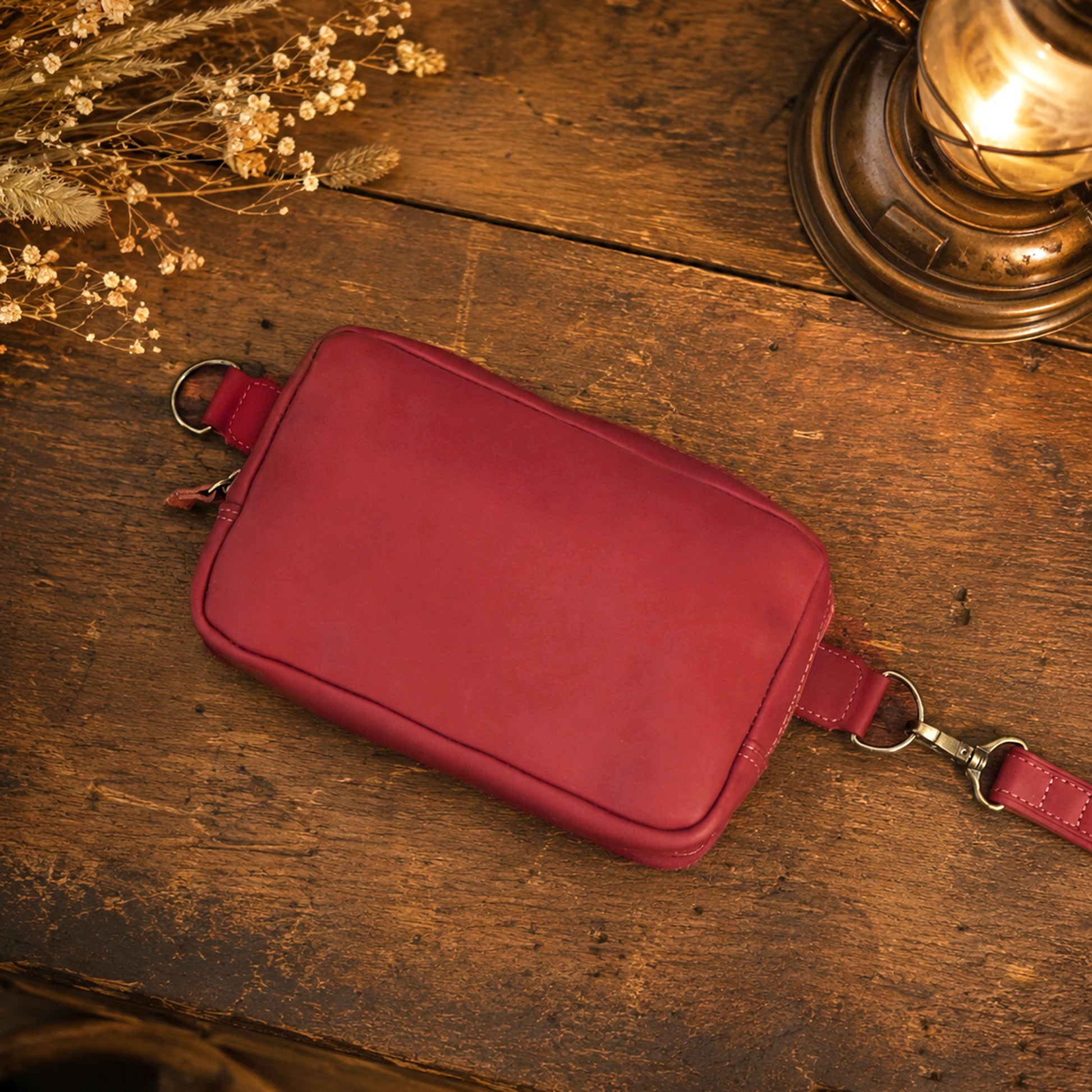 A rectangular red leather crossbody bag with a matching strap and silver-tone hardware, displayed on a rustic wooden surface with dried flowers and a brass lamp in the background.