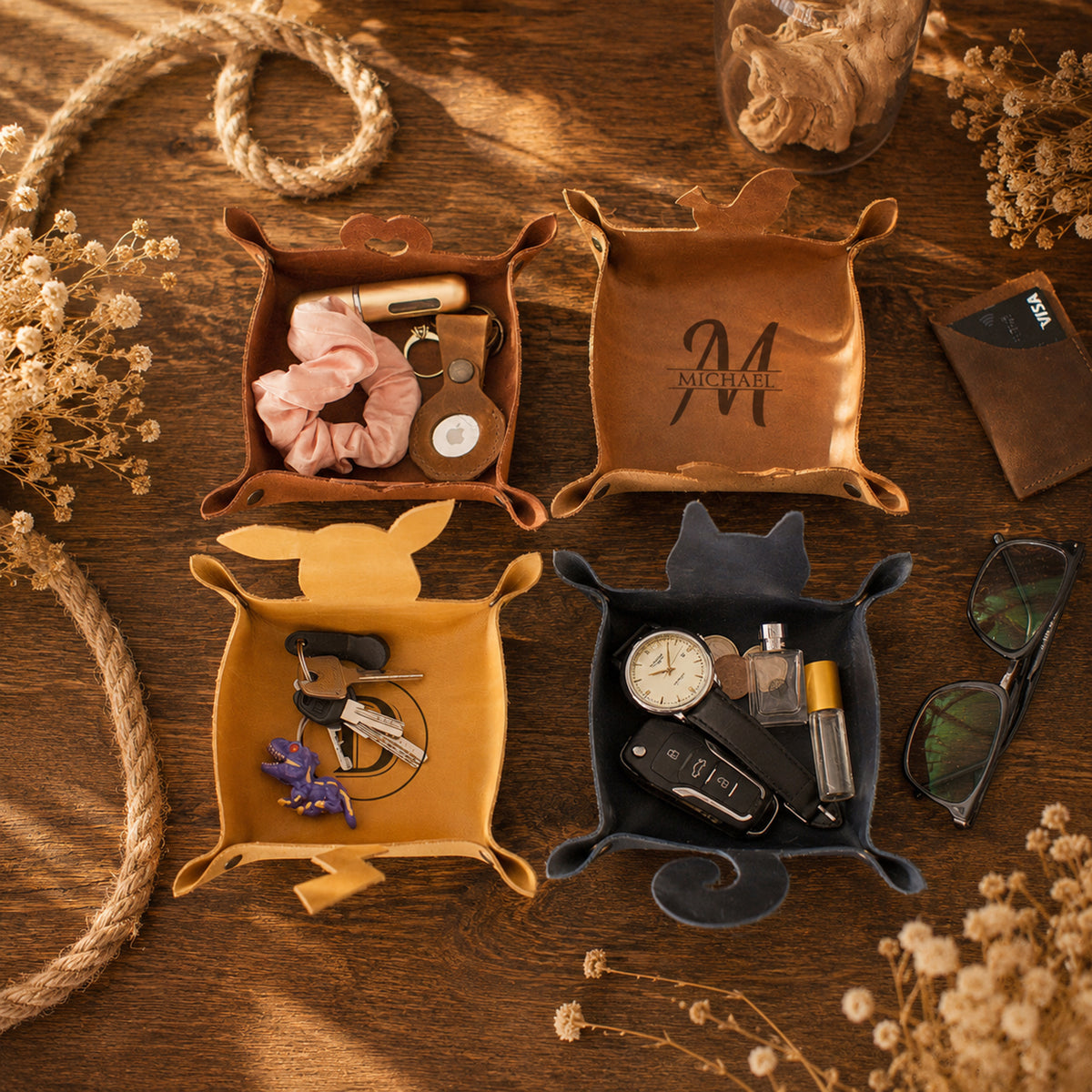 Four leather key holder trays in brown, tan, mustard yellow, and charcoal gray, arranged on a wooden table with dried flowers, rope, and other accessories. The top-left tray holds a pink scrunchie and a small bag, the top-right tray is open with a monogram 'M' on the lid, the bottom-left tray is open with keys and a purple flower, and the bottom-right tray holds a car key fob, perfume bottle, and sunglasses.