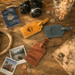 Three personalized leather luggage tags in tan, brown, and blue-gray are arranged on a rustic wooden table with travel-themed props including a passport, camera, coins, a map, and instant photos.