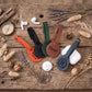 Four personalized leather AirTag keychain phone straps in orange, black, green, and brown, styled on a rustic wooden table with decorative dried flowers, a compass, and white earbuds.