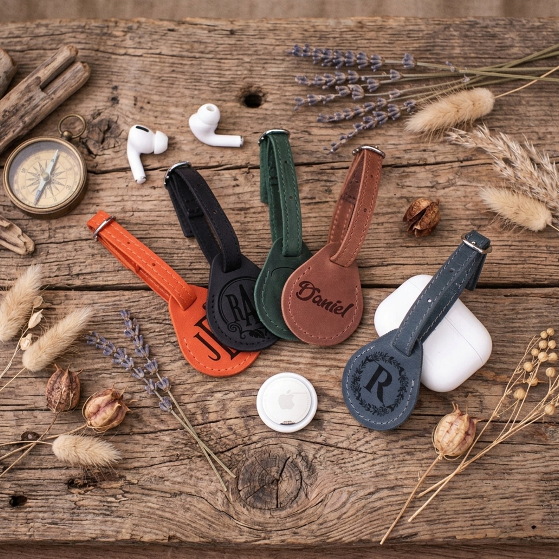 Four personalized leather AirTag keychain phone straps in orange, black, green, and brown, styled on a rustic wooden table with decorative dried flowers, a compass, and white earbuds.