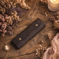 A dark brown leather jewelry organizer case with snap buttons, styled on a rustic wooden table with dried flowers, necklaces, rings, and a lit candle.