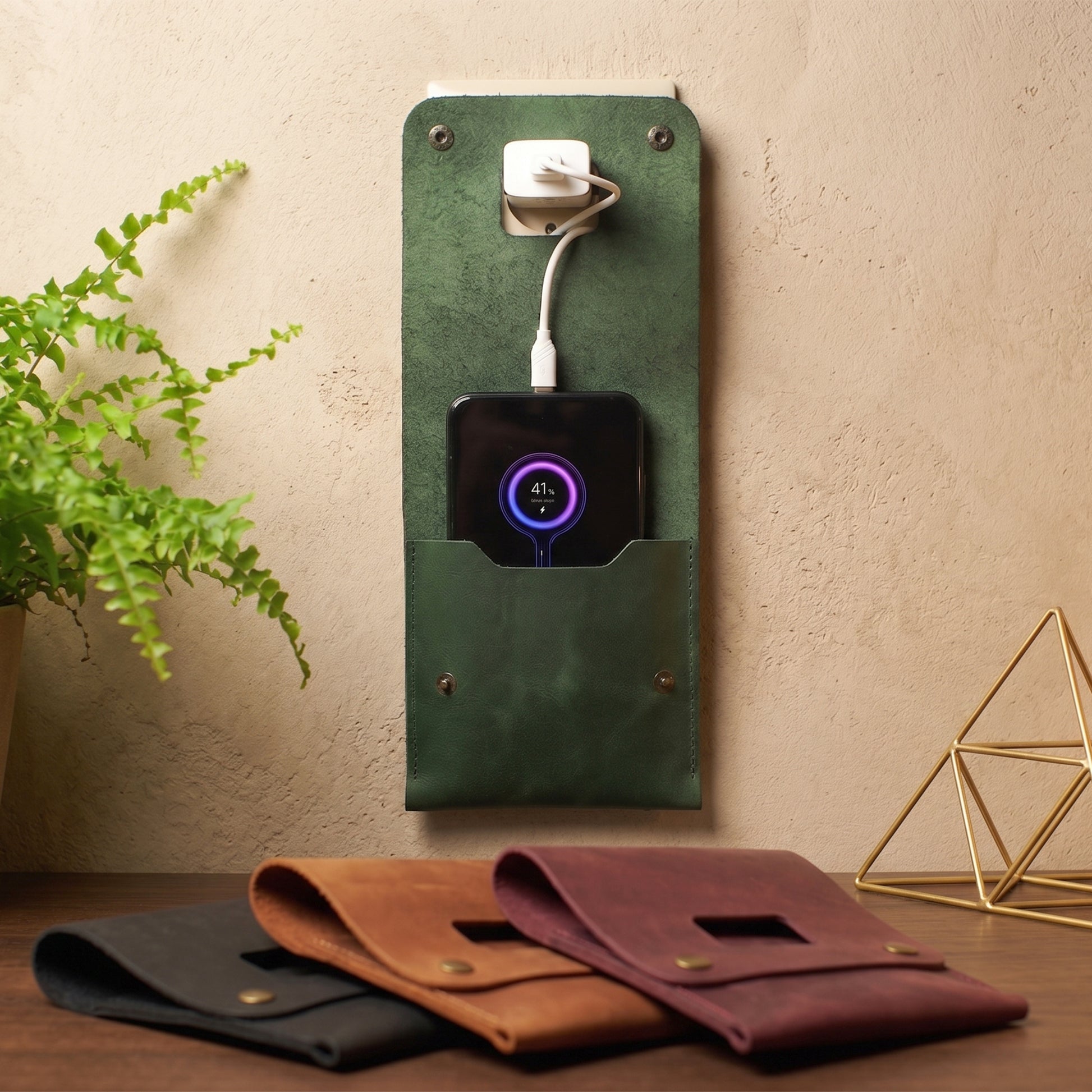 A dark green leather wall-mounted charging stand holding a black smartphone with a charging cable plugged into the wall, set against a beige wall. In the foreground, three matching leather pouches in black, brown, and burgundy rest on a wooden surface next to a green plant and a gold geometric decor piece.