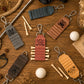 Six personalized leather golf keychains arranged in a flat-lay on a wooden table with golf tees, golf balls, and rustic props. The keychains are in various colors (gray, teal, brown, dusty red, tan, and dark brown) and each features a unique engraved design, including a monogram 'M', a golf cart, a duck, a golf ball with a crown, and a golf flag.