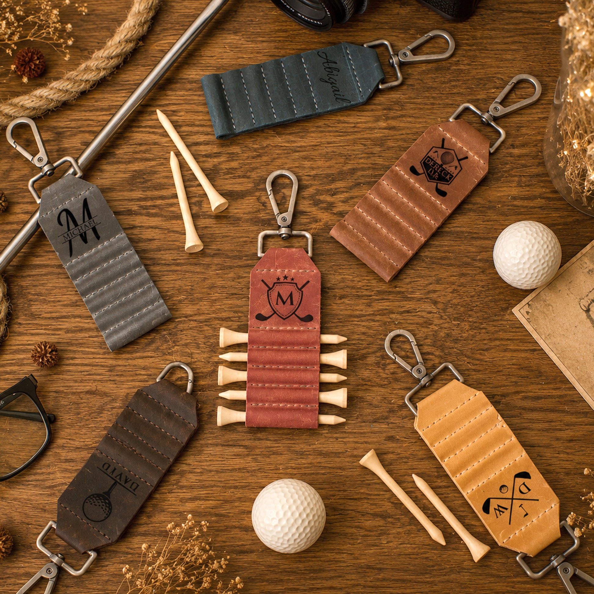 Six personalized leather golf keychains arranged in a flat-lay on a wooden table with golf tees, golf balls, and rustic props. The keychains are in various colors (gray, teal, brown, dusty red, tan, and dark brown) and each features a unique engraved design, including a monogram 'M', a golf cart, a duck, a golf ball with a crown, and a golf flag.