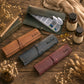 Three personalized leather pill organizer rolls in brown, blue-gray, and burgundy, arranged on a rustic wooden table with dried flowers, books, capsules, and supplement bottles.