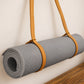 A gray textured yoga mat rolled up and held by a tan leather strap, shown against a plain off-white wall with a wooden surface at the bottom.
