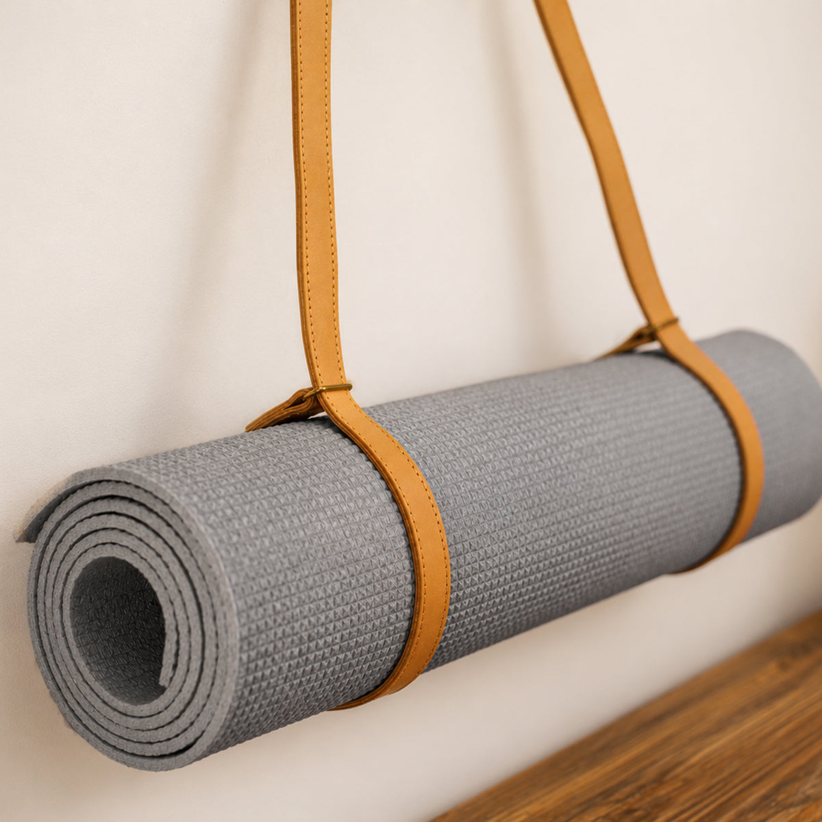 A gray textured yoga mat rolled up and held by a tan leather strap, shown against a plain off-white wall with a wooden surface at the bottom.