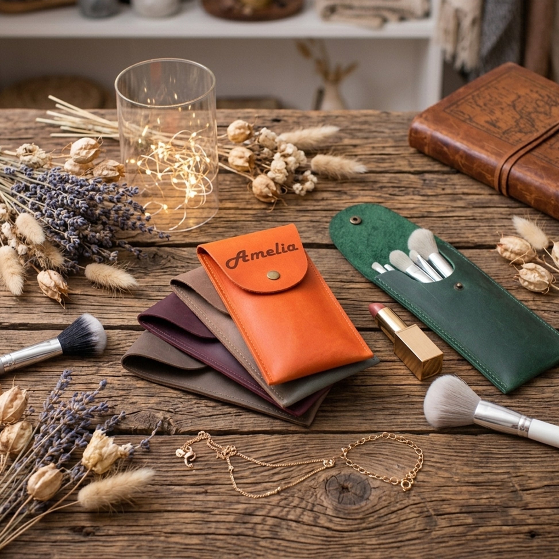 A styled flat-lay of personalized makeup pouches on a rustic wooden table, surrounded by dried lavender, a glass candle holder with fairy lights, brushes, and a notebook. The central product is an orange leather pouch personalized with the name 'Amelia' in black script.