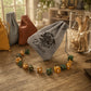 A gray personalized drawstring pouch with 'Dave' and a rose emblem printed on it, displayed on a wooden table next to a bracelet of multicolor polyhedral dice. Other pouches and a glass jar are blurred in the background.