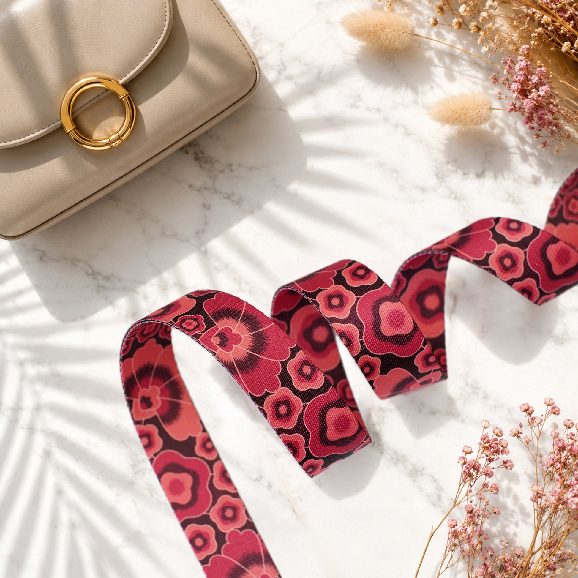 Beige handbag with a red floral strap on a marble surface with dried flowers.