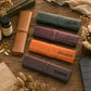 Four personalized leather travel passport covers in brown, green, orange, and purple, arranged on a rustic wooden table with vintage props like a map, dried flowers, and a pill bottle.