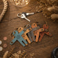 A set of personalized leather keychains shaped like stylized letters, displayed on a rustic wooden table with decorative props including a rope, dried flowers, and a key.