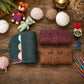 Two personalized leather zippered cases, one dark green and one brown, displayed on a rustic wooden table surrounded by knitting and crochet props including yarn balls, a small plush bear, knitting needles, and a crochet hook.