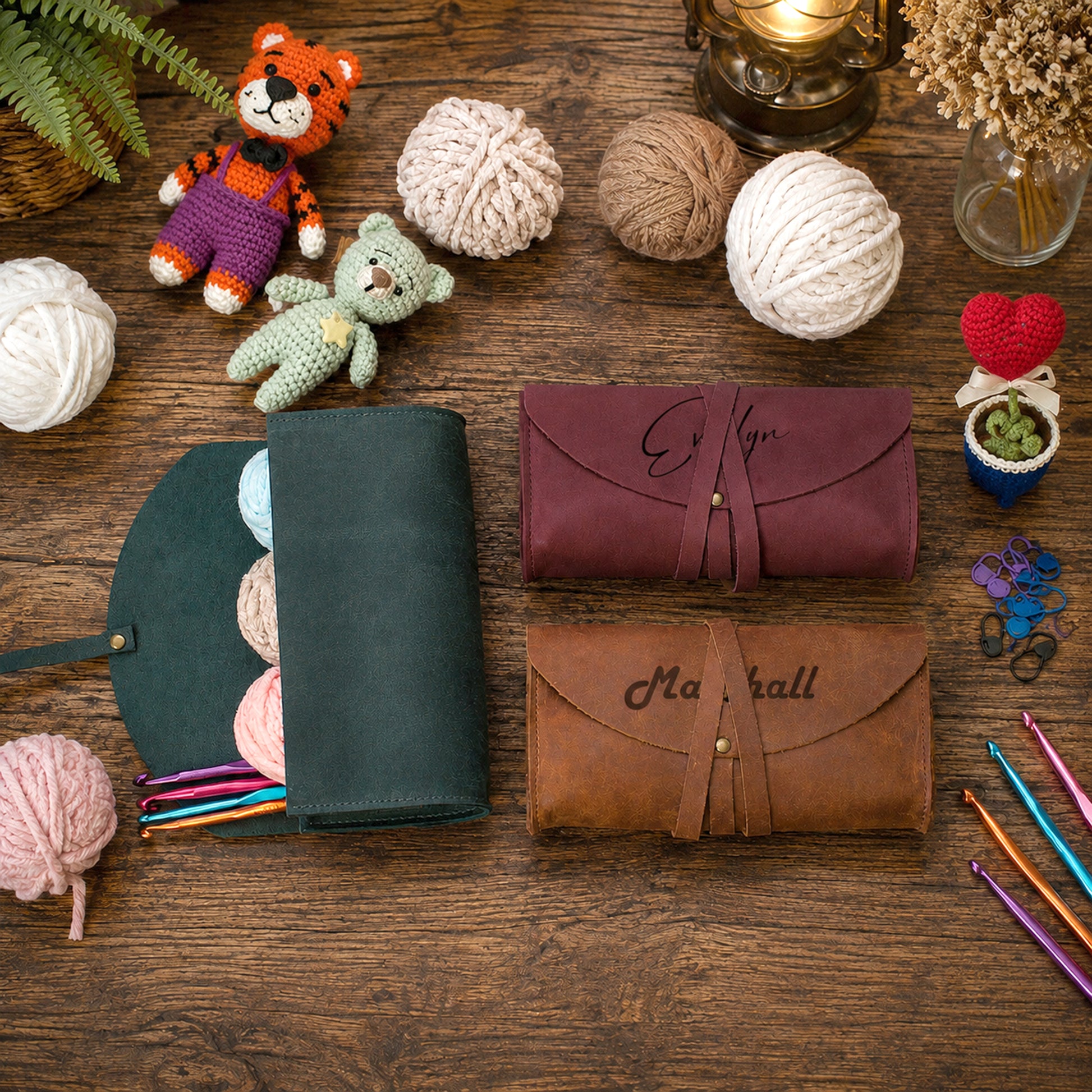 Two personalized leather zippered cases, one dark green and one brown, displayed on a rustic wooden table surrounded by knitting and crochet props including yarn balls, a small plush bear, knitting needles, and a crochet hook.