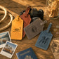 Three personalized leather luggage tags in yellow, brown, and blue-gray are arranged on a rustic wooden table with a Nikon camera, rope, coins, Polaroid photos, and a passport. The tags are engraved with outdoor scenes and names.