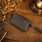 A black leather wristlet pouch with a strap and metal rings, styled on a rustic wooden table with dried flowers, a vintage book, and a glowing brass lantern.