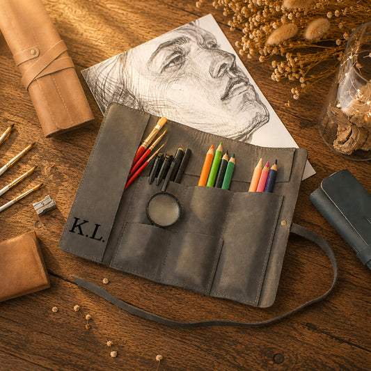 A dark gray leather artist's roll-up pouch laid open on a wooden table, holding colored pencils, brushes, and pens. A sketch of a face is placed behind it, with dried flowers, a rolled leather sleeve, and other art supplies around it.
