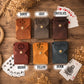 Six leather card envelopes in different colors (Tawny, Gray, Chocolate, Brown, Yellow, and Denim) arranged in a 2x3 grid on a rustic wooden background with playing cards and dried flowers. Each envelope has a different engraved name or logo and a gold snap button.