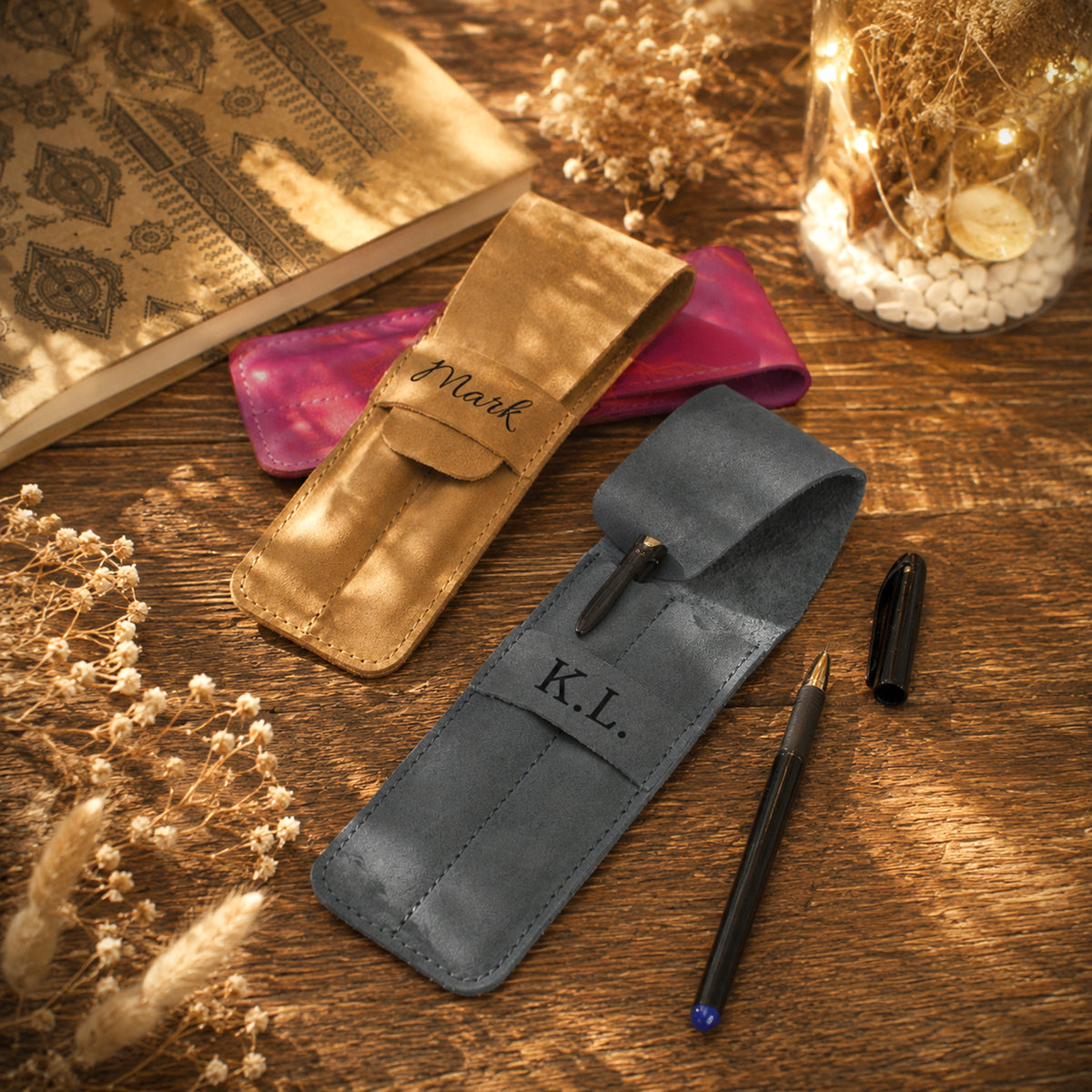 Two personalized leather pen cases on a rustic wooden table, one in tan with the name 'Mark' and one in slate blue with 'K.L.