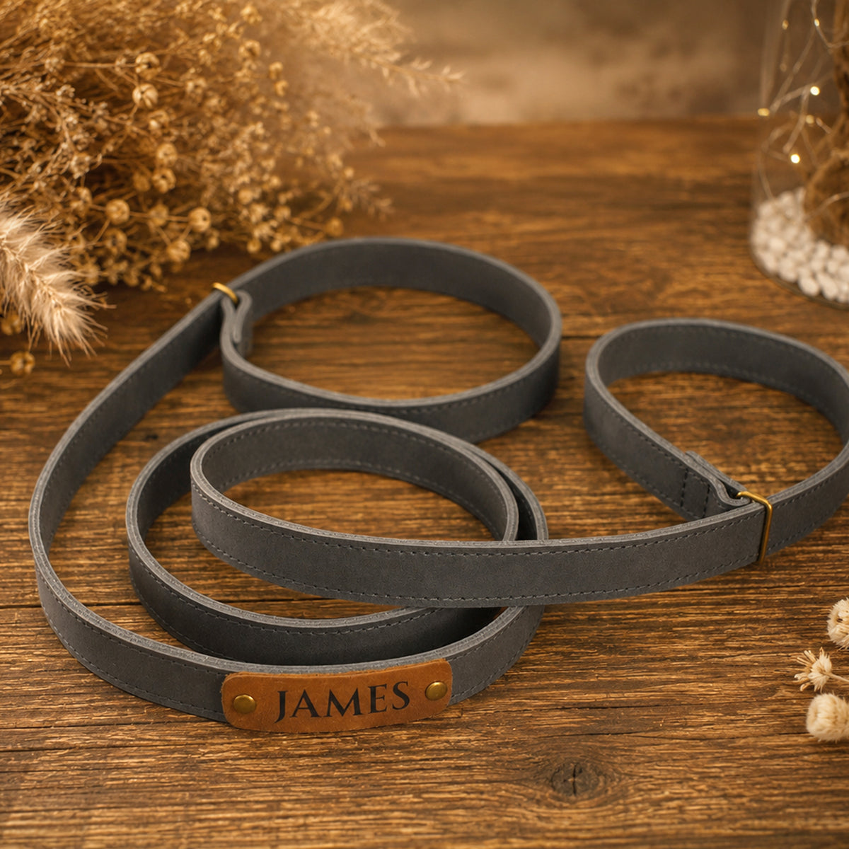 A dark gray faux-leather dog leash with a brown leather nameplate engraved with 'JAMES' and gold-tone metal studs, displayed on a rustic wooden surface with dried floral decor.
