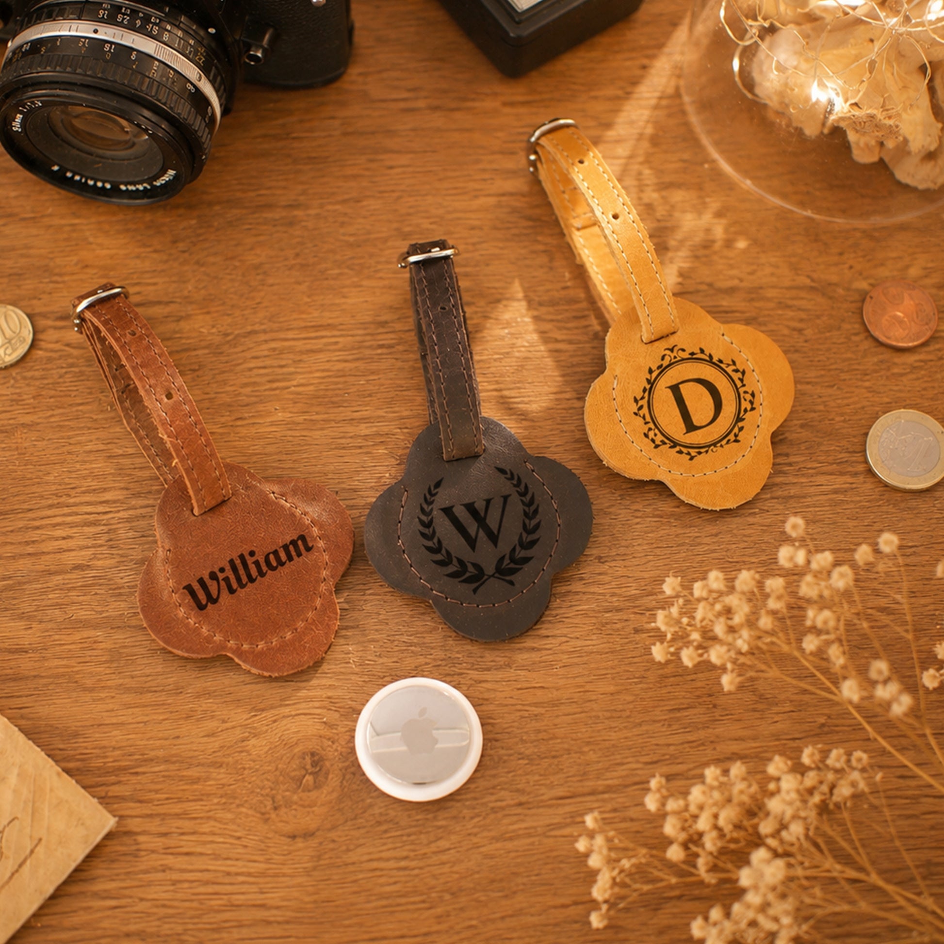 Three personalized leather luggage tags with cloud-shaped metal plates are arranged on a wooden tabletop. One tag is light brown with the name 'William', one is dark brown with a laurel wreath and 'W', and one is tan with a decorative monogram 'D'.