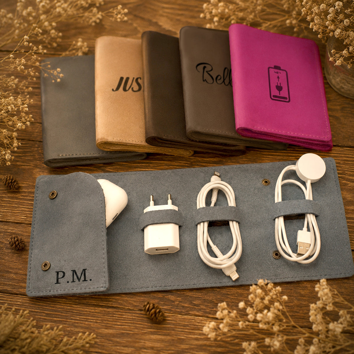 A personalized gray faux-leather roll-up organizer laid on a wooden surface, holding a white charging cable, white wall charger, and a white charging puck. Behind it are four matching wallet-style cases in gray, tan, brown, and magenta, each with engraved text or a battery icon.
