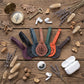 Five leather earbud strap protectors in different colors (purple, black, brown, green, orange) are arranged on a rustic wooden background with styled flat-lay props including a brass compass, vintage key, dried lavender, and white wireless earbuds and charging cases.