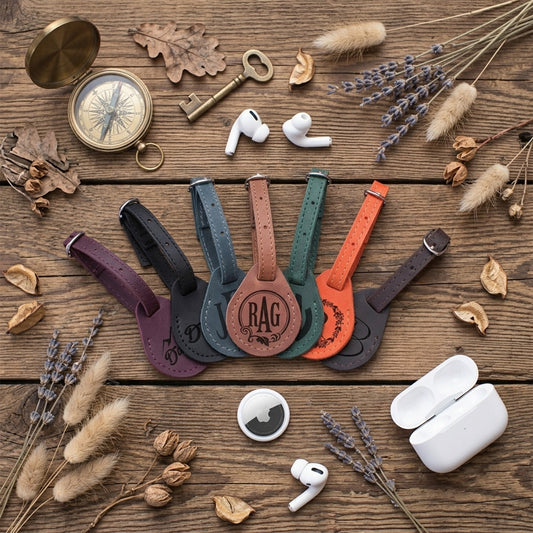 Five leather earbud strap protectors in different colors (purple, black, brown, green, orange) are arranged on a rustic wooden background with styled flat-lay props including a brass compass, vintage key, dried lavender, and white wireless earbuds and charging cases.