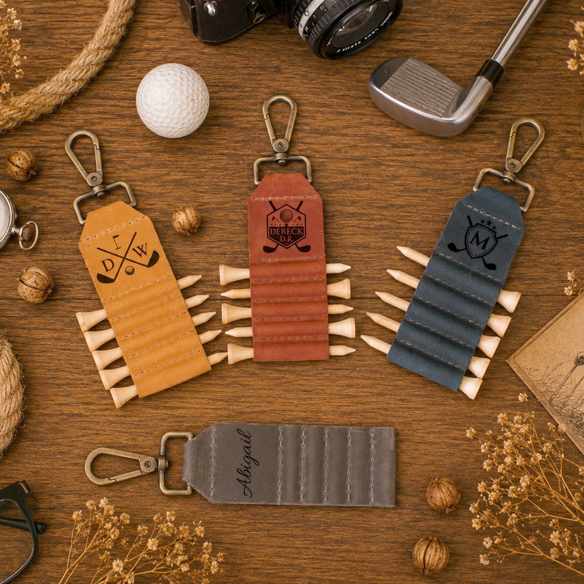 Three golf-themed keychains with wooden spike-like accents are arranged on a wooden tabletop with golf props and dried flowers. The keychains are shown in tan, reddish-brown, and slate blue, with one additional gray personalized keychain below.
