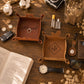 Two personalized brown leather tray desk organizers on a wooden table, surrounded by lifestyle props including an open book, watch, sunglasses, keys, and cotton bolls. One tray is dark brown with the name 'Abigail' and a ring inside, and the other is tan with 'AJF' inside a diamond.