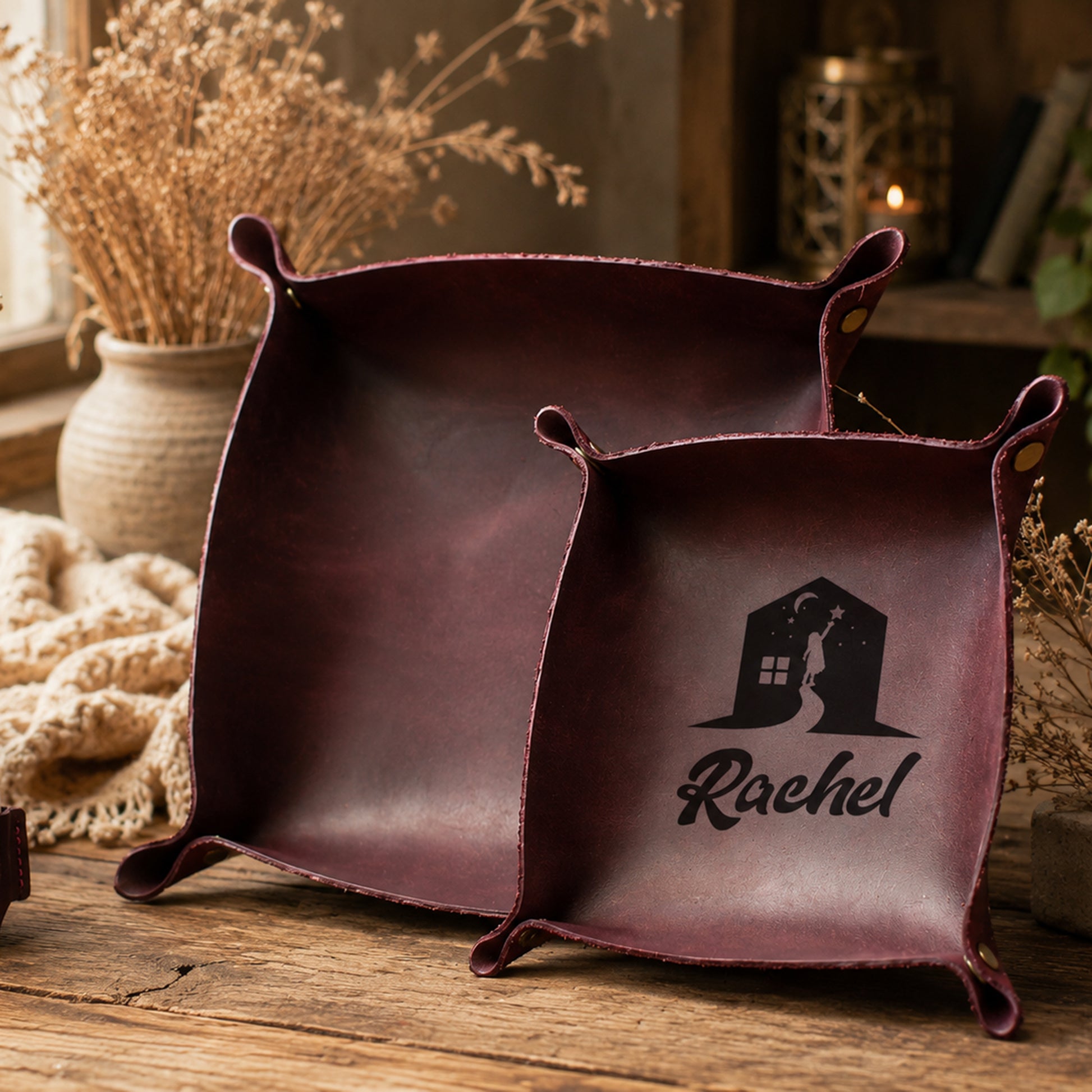 Two square dark burgundy leather storage bins with handles, one plain and one personalized with a black house silhouette and the name Rachel, displayed on a rustic wooden table with dried flowers and a cozy background.