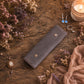 A dark gray velvet jewelry organizer case with snap buttons, styled on a rustic wooden table surrounded by dried flowers, a lit candle, rings, and a necklace.