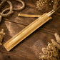 A tan leather drumstick sleeve holding two light wood drumsticks, arranged diagonally on a rustic dark wood tabletop with dried flowers and rope as styled props.